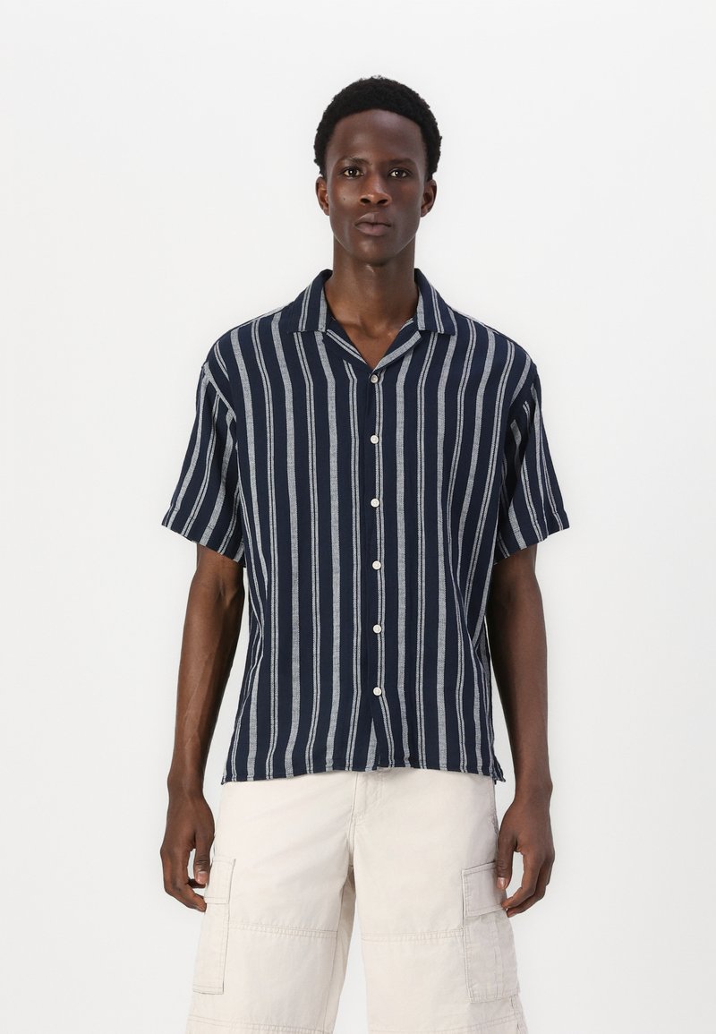 Chemise à rayures bleu marine et blanche à manches courtes avec col, fermeture à boutons et coupe décontractée. Portée avec un short cargo de couleur claire.