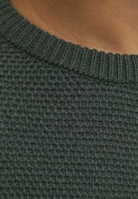 Nära bild av mörkgrön stickad tröjtyg med ribbad krage runt en persons hals och övre bröst.