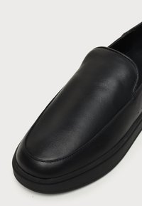 Μαύρο δερμάτινο παπούτσι τύπου slip-on με στρογγυλεμένη τετράγωνη μύτη και παχιά σόλα σε απλό φωτεινό φόντο.