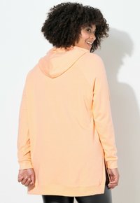 Mujer con cabello rizado vestida con sudadera con capucha melocotón, sonriendo mientras mira por encima de su hombro contra un fondo blanco.