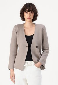 Lys beige cardigan laget av teksturert stoff, med en enkelt svart knapp, åpen front og lange ermer. Bært over en svart topp.