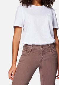Camiseta blanca de manga corta con un bolsillo, combinada con jeans marrones claros que tienen bolsillos frontales curvados y un diseño ajustado.