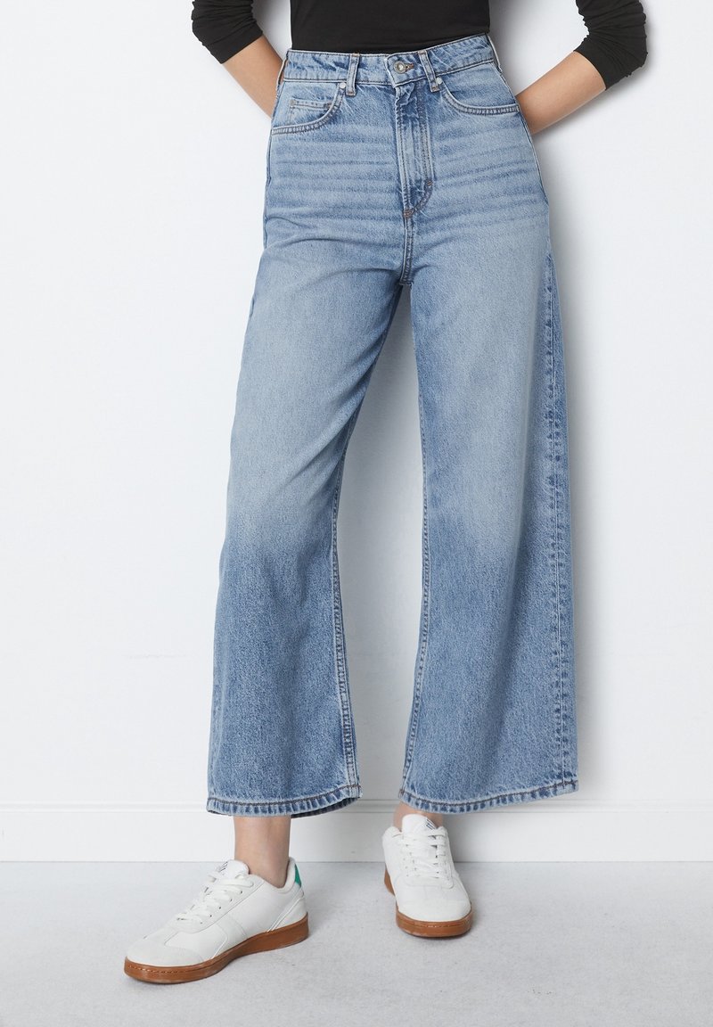 Personne porte un jean bleu clair à jambes larges et des baskets blanches, debout contre un mur blanc uni les mains dans les poches.