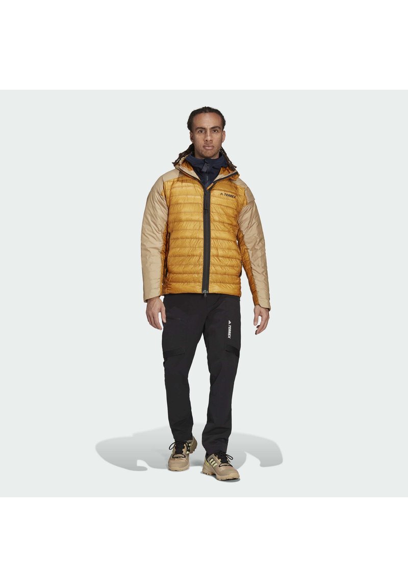 Gelbe und beigefarbene isolierte Jacke mit horizontalen Nähten, schwarze Hose und hellbraune Schuhe. Verfügt über eine Kapuze und einen Reißverschluss vorne.