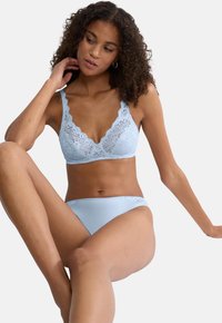 Ženska s kodrasto pričesko, oblečena v svetlo modro čipkasto modrček in usklajene hlačke, sedi s povzdignjenim kolenom na belem ozadju.