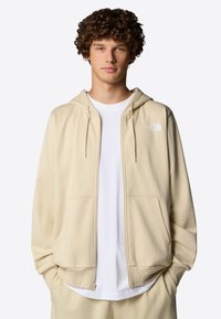 Hoodie zippé beige en tissu doux, avec un logo à l'avant, deux poches latérales et une capuche avec cordon de serrage. Porté sur une chemise blanche.