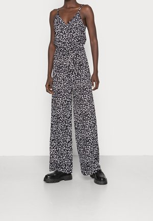 Kvinde iført en sort ærmeløs jumpsuit med hvide abstrakte prikker, vide ben, et bindebælte i taljen og sorte kraftige sko.