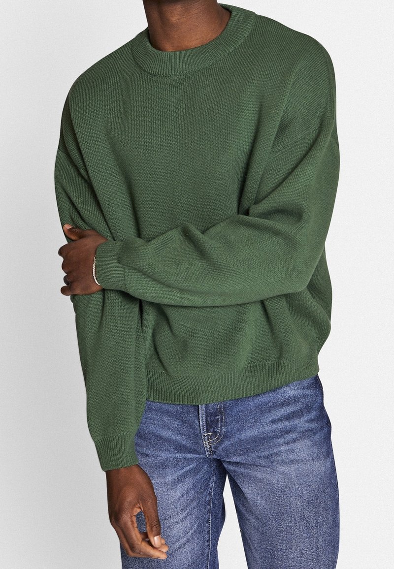 Personne portant un pull tricoté vert ample et un jean bleu, avec un bras croisé sur la poitrine, sur un fond uni.
