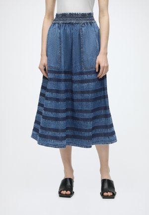 Donna che indossa una gonna midi in denim blu con righe orizzontali, grandi tasche frontali e scarpe nere aperte con tacco quadrato.