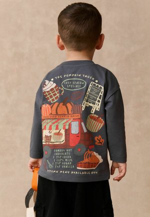 Niño con una camiseta de manga larga oscura con un camión de calabazas con tema otoñal, receta de chocolate caliente e ilustraciones estacionales en la parte trasera.