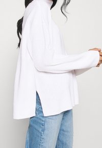Weißer Rollkragenpullover mit gerippter Textur, lockerer Passform und seitlichen Schlitzen, kombiniert mit hellblauen Jeans.