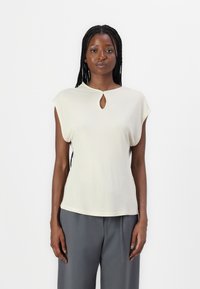 Haut court beige clair à manches courtes avec un large col et un petit détail en forme de trou de clé au centre. Texture lisse, coupe décontractée.