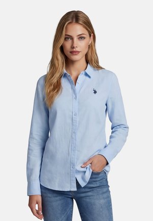 Woman with long blonde hair wearing light blue button-up shirt with logo on chest and blue jeans, standing with one hand in pocket.