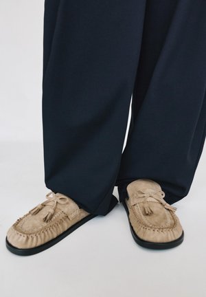 Beżowe zamszowe mokasyny z frędzlami noszone z czarnymi spodniami o szerokich nogawkach na białym tle.