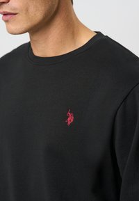 Hombre vistiendo una camiseta de cuello redondo negra con un pequeño logo de un jugador de polo bordado en rojo en el lado izquierdo del pecho.
