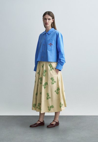 Γυναίκα που φοράει μπλε πουκάμισο με κουμπιά και κεντημένο λογότυπο και μπεζ φούστα με πράσινα λουλουδάτα σχέδια, στέκεται με τα χέρια στις τσέπες της φούστας.