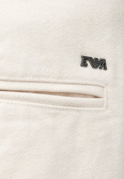 Hvit jakke med strukturert stoff, med en horisontal lomme og en sølvlogo på øvre venstre side.