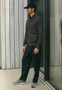 Grauer Reißverschluss-Pullover mit strukturiertem Design, dunklen Hosen und grauen Sneakern, ergänzt durch eine marineblaue Mütze. Schlicht, entspannter Schnitt und moderner Stil.
