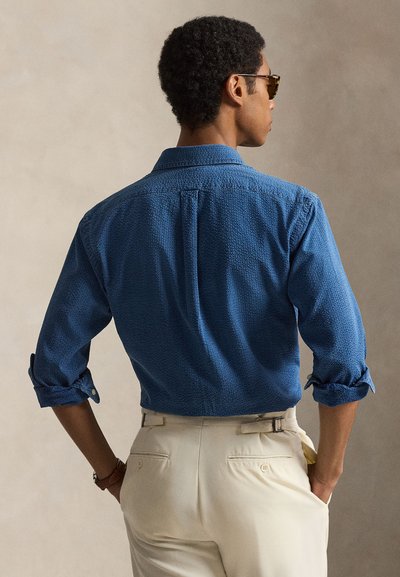 Mees kannab tekstuuriga sinist pikkade varrukatega särki, mille varrukad on üles rullitud, ja beeži värvi pükse, seisab selg inimese poole, käed taskutes ja päikeseprillid peas.