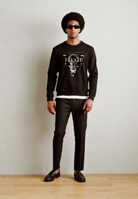 Sudadera negra con un gráfico de un pantera, combinada con pantalones negros a medida y mocasines oscuros. El modelo lleva gafas de sol y está de pie frente a un fondo sencillo.