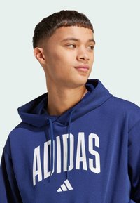 Blå huvtröja med vit "ADIDAS"-logotyp. Har dragsnören och mjukt tyg. Enkel design utan ytterligare mönster eller detaljer.