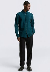 Säästvalt tekstuuritud kangast türkiissinine nööbitav särk, mis on kombineeritud mustade pükste ja mustade saabastega. Särgil on krae ja esikinnis nööpidega.