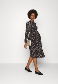 Vestido midi preto com flores e mangas compridas, apresentando padrões em rosa e verde. Combinado com mocassins negros e uma bolsa prateada texturizada.