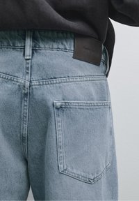 Gros plan de la poche arrière d'un jean denim bleu clair avec boucle de ceinture visible et étiquette de marque en cuir noir.