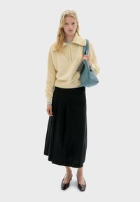 Junge Frau mit langen blonden Haaren, die einen cremefarbenen Strickpullover, einen schwarzen Midirock, schwarze Absätze trägt und eine blaue Umhängetasche hält.