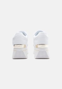 Un paio di sneakers bianche con piattaforma e dettagli dorati, con il logo "GUESS" sul tallone, viste da dietro su uno sfondo neutro.