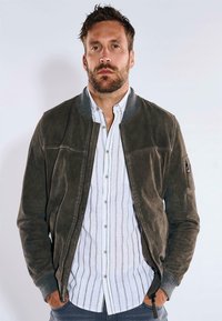Dunkelgrüne Leder-Bomberjacke mit geripptem Kragen, Reißverschluss vorne und Seitentaschen, getragen über einem weiß gestreiften Hemd.