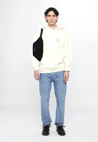 Cream hoodie with front pocket and logo, blue jeans, and black shoes. Black sling bag worn on the left side. Model stands against a white background.
