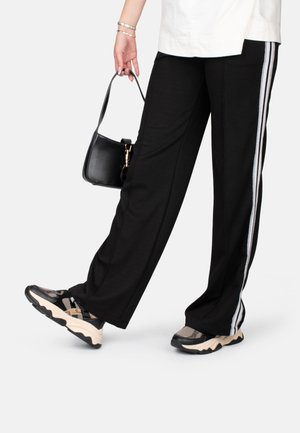 Personne portant un pantalon noir à jambes larges avec des bandes blanches sur les côtés, des baskets noires et beiges, un haut blanc et tenant un petit sac à main noir.