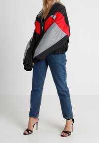 Giubbotto bomber nero con blocchi di colore rosso e grigio, chiusura con cerniera e polsini elasticizzati. Abbinato a jeans a vita alta blu e sandali neri.