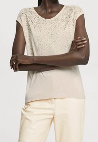Krátký béžový top s kulatým výstřihem, zdobený texturovaným designem kovových teček na přední straně. Hladká, měkká tkanina.