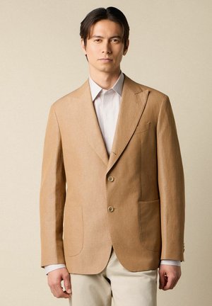 Man draagt een beige blazer over een gestreept wit shirt en lichte broek, staand tegen een effen beige achtergrond.