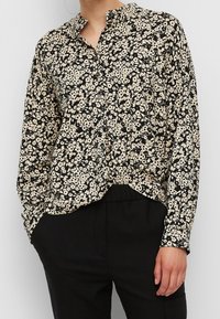 Blouse boutonnée à manches longues, noire et beige avec motif floral, portée non rentrée sur un pantalon noir, une main dans la poche.