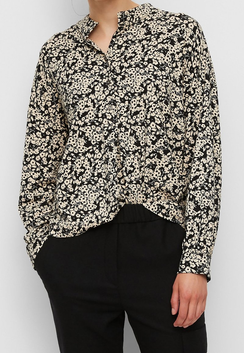 Blouse boutonnée à manches longues, noire et beige avec motif floral, portée non rentrée sur un pantalon noir, une main dans la poche.