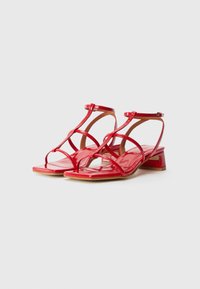 Red patent leather heels with a block heel, featuring crisscross straps and an ankle buckle. Square toe design with smooth finish.