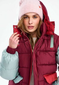 Nő, aki egy bordó és világoskék pufidzseki kapucniját igazítja, rózsaszín kötött sapkát és bézs felsőt visel, előre néz neutrális arckifejezéssel.