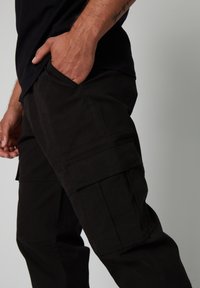 Zwarte cargobroek van stevig materiaal heeft meerdere zijzakken, een rechte pasvorm en een gladde, matte textuur.