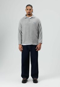 Grijze fleece trui met een kraag en een blauw logo, gecombineerd met donkerblauwe broek met brede pijpen. Zwarte schoenen met gekleurde veters maken de outfit compleet.