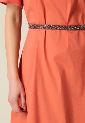Frau, die ein korallenfarbenes Kleid mit kurzen Ärmeln trägt, in der Taille durch einen mit Perlen besetzten, funkelnden Gürtel betont.