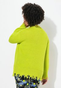 Un pull en tricot vert citron à coupe décontractée, avec un ourlet effiloché et un tissu texturé, porté sur un pantalon camouflage à motifs.