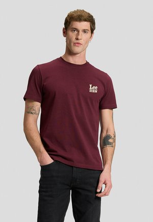 Hombre con camiseta Lee Denim burdeos y vaqueros negros, de pie con una mano en el bolsillo sobre un fondo liso gris claro.