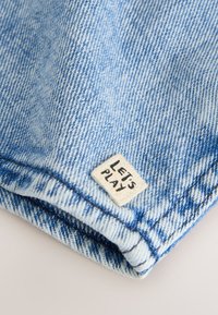 Nahaufnahme von blauer Denim-Stoff mit einer gefalteten Kante und einem kleinen weißen Etikett, auf dem "LET'S PLAY" in schwarzen Buchstaben steht.