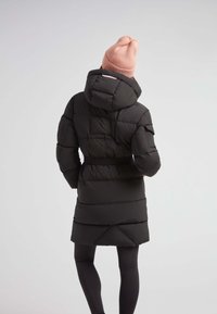 Abrigo acolchado negro con capucha, cintura ceñida con cinturón y hombros texturizados. Combinado con un gorro de punto rosa claro. Diseño simple sin logotipos visibles.