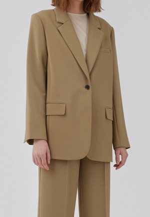 Kobieta w beżowej, oversize marynarce z klapami z wcięciem, czarnym guzikiem, kieszeniami z patkami oraz pasujących szerokich spodniach na jasnej bluzce.