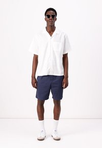 White short-sleeve shirt with a pocket, navy shorts, white socks, and beige sneakers. Accessorized with dark sunglasses.