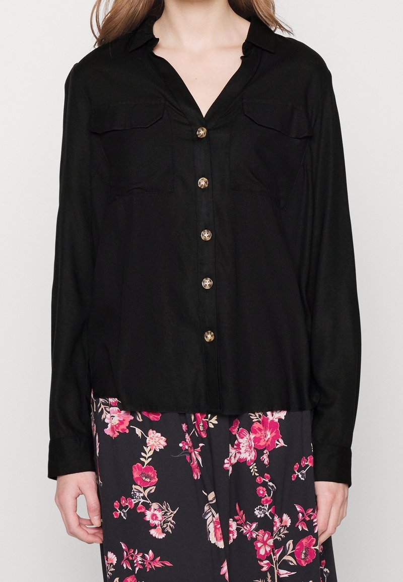 Chemise noire à col, avec deux poches poitrine et cinq boutons de couleur claire. Manches longues avec poignets à boutons et texture de tissu lisse.